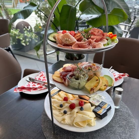 Eine dreistöckige Präsentation mit Aufschnitt, Käse, Obst und einer Topfpflanze. Teller mit Besteck und Gewürzen stehen auf dem Tisch.