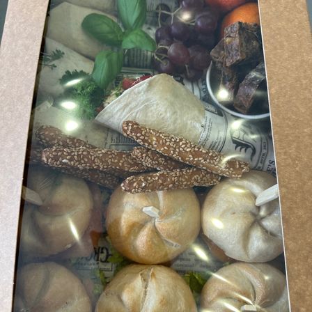 Eine Schachtel zeigt eine Auswahl an Lebensmitteln, darunter Brötchen, Sesam-bedeckte Brotstangen, Trauben, getrocknete Früchte und Kräuter. Die Schachtel ist geöffnet und zeigt eine Zeitung mit einem Bild eines Löwen darin.