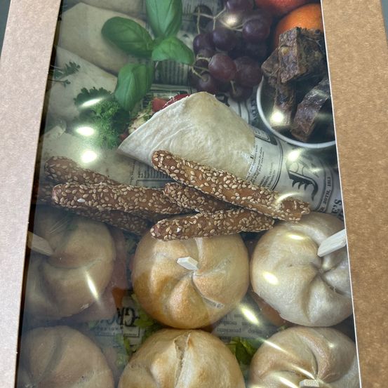 Eine Schachtel zeigt eine Auswahl an Lebensmitteln, darunter Brötchen, Sesam-bedeckte Brotstangen, Trauben, getrocknete Früchte und Kräuter. Die Schachtel ist geöffnet und zeigt eine Zeitung mit einem Bild eines Löwen darin.