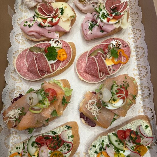 Eine Schachtel mit Sandwiches mit verschiedenen Belägen wie Käse, Fleisch, Gemüse und Kräutern, auf einem Spitzentuch angeordnet.