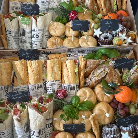 Eine Auswahl an Sandwiches und Gebäck wird in einer Box präsentiert, darunter Wraps, Baguettes und Sandwiches. Es gibt auch Obst und Gemüse zur Schau gestellt.
