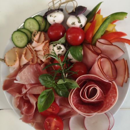 Ein weißer Teller zeigt eine Auswahl an Aufschnitt, geschnittenen Gurken, Kirschtomaten und bunten Gemüsesorten. Mozzarella-Kugeln mit Kräutern und Basilikumblättern sind ebenfalls auf dem Teller arrangiert.