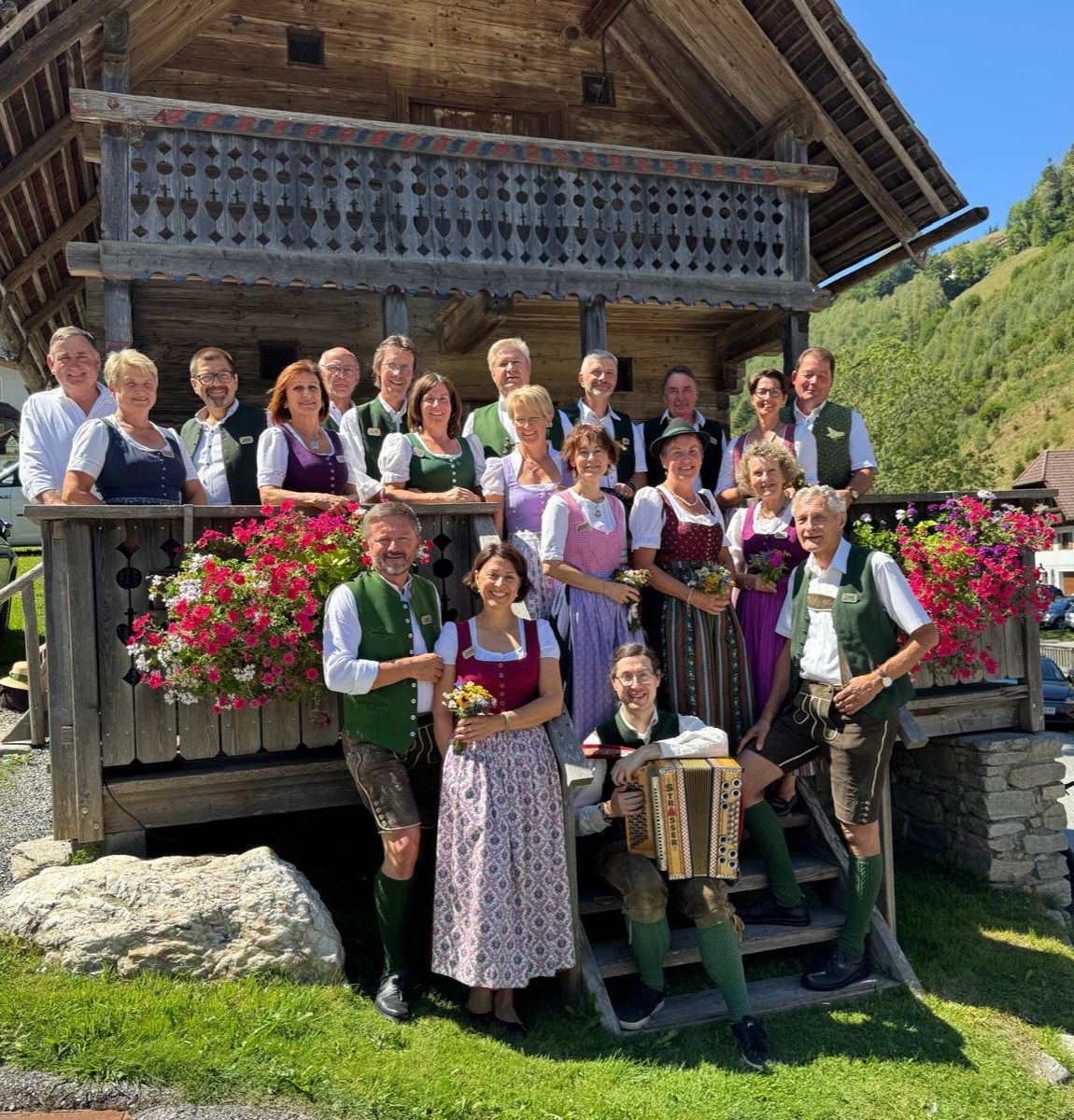 Eine Gruppe von Menschen in traditioneller Kleidung posiert für ein Foto vor einem Holzhaus mit Balkon und Blumen.