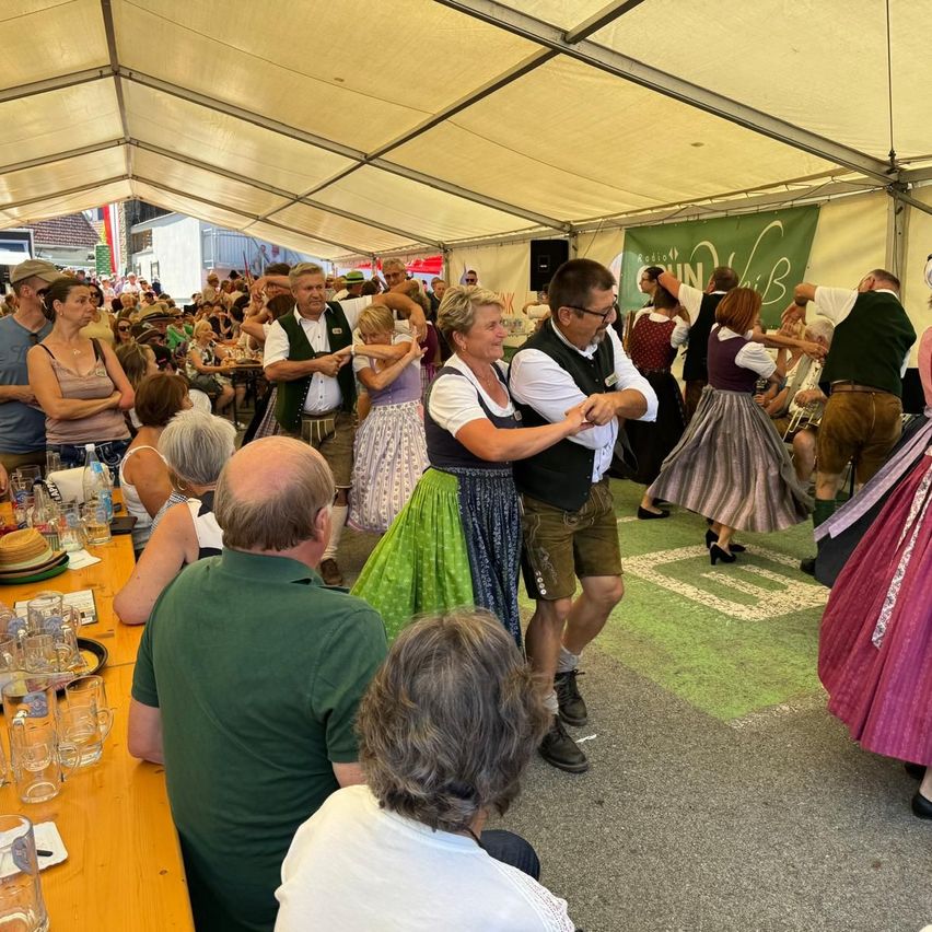Eine Gruppe von Menschen tanzt in einem Zelt mit weißer Decke und grüner Fahne. Einige tragen traditionelle Kleidung.