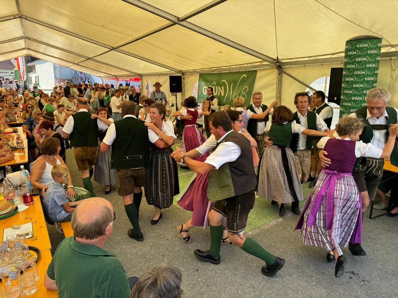 Eine Gruppe von Menschen in traditioneller bayerischer Kleidung tanzt im Kreis unter einem Zelt. Ein Banner mit Text ist im Hintergrund sichtbar.