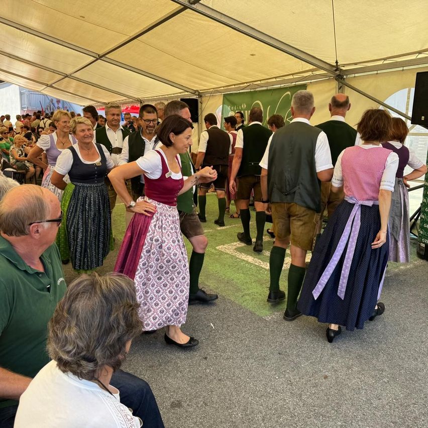 Eine Gruppe von Menschen in traditioneller deutscher Kleidung tanzt in einem Zelt bei einer Veranstaltung. Einige sitzen, während andere stehen und tanzen.