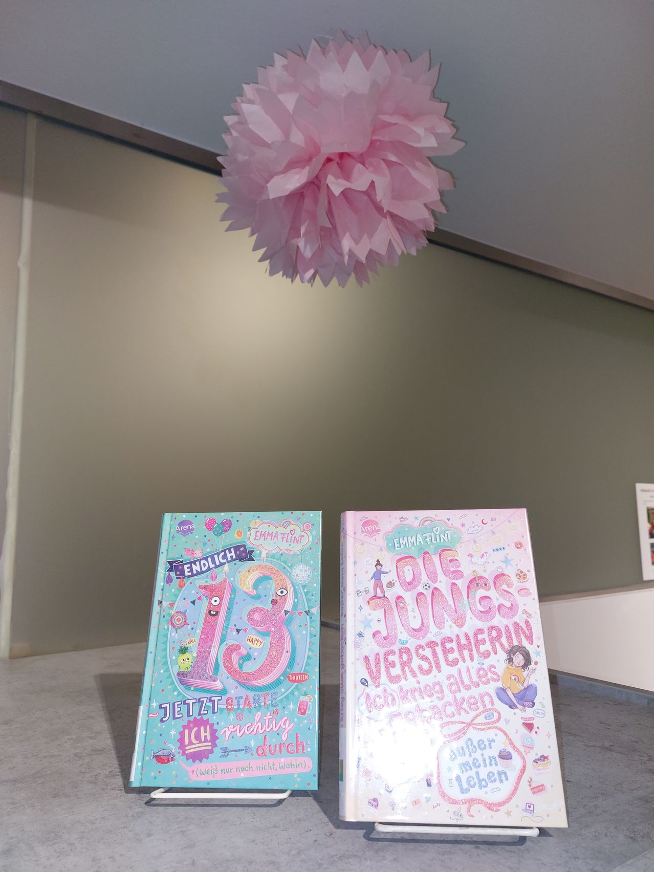 Zwei Bücher sind auf einem Tisch ausgestellt. Eines heißt 'Endlich 13' und das andere 'Die junge versteherin'. Über den Büchern hängt eine rosa Papierdekoration von der Decke. Der Hintergrund ist eine beige Wand.