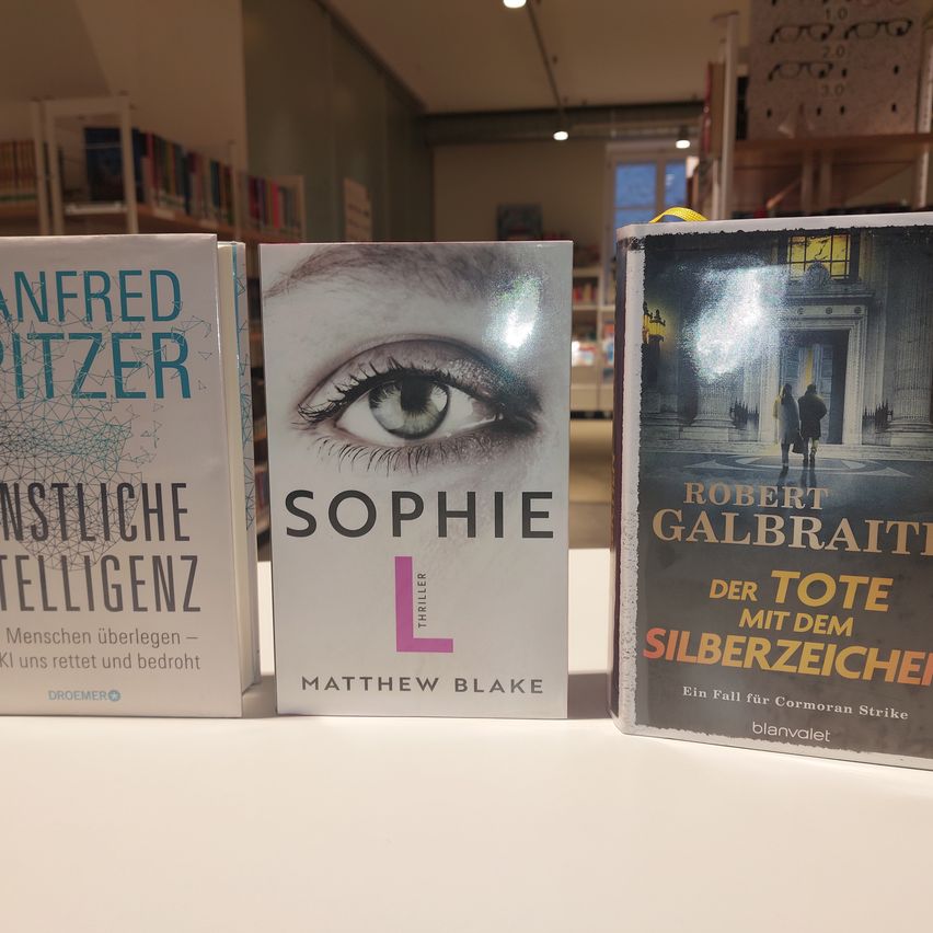 Drei Bücher stehen auf einem Tisch. Ein Buch von Matthew Blake heißt 'Sophie L'. Ein weiteres von Robert Galbraith heißt 'Der Tote mit dem Silberzeichen'. Das dritte Buch ist von Manfred Spitzer.