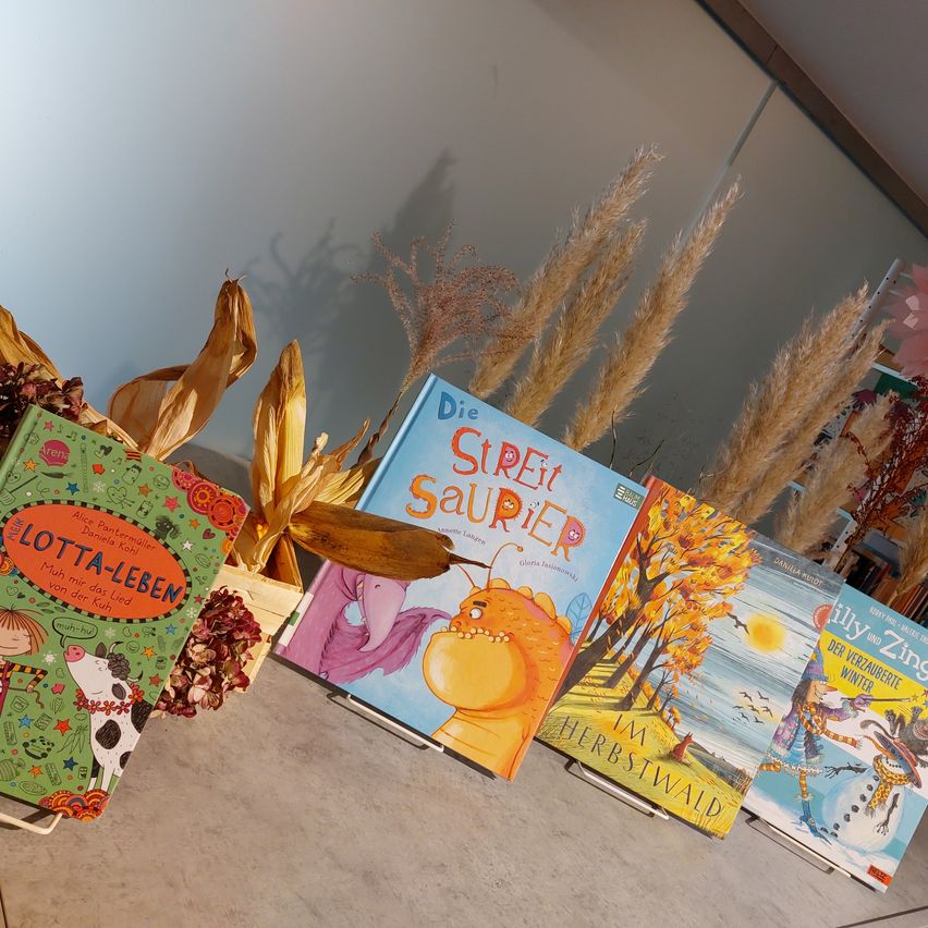 Eine Sammlung von Kinderbüchern ist auf einem Tisch ausgestellt, mit einer weißen Wand im Hintergrund. Die Bücher umfassen 'Lotta-Leben', 'Die Sirei Saurier', 'Im Herbstwald' und 'Zilly und Zing'.