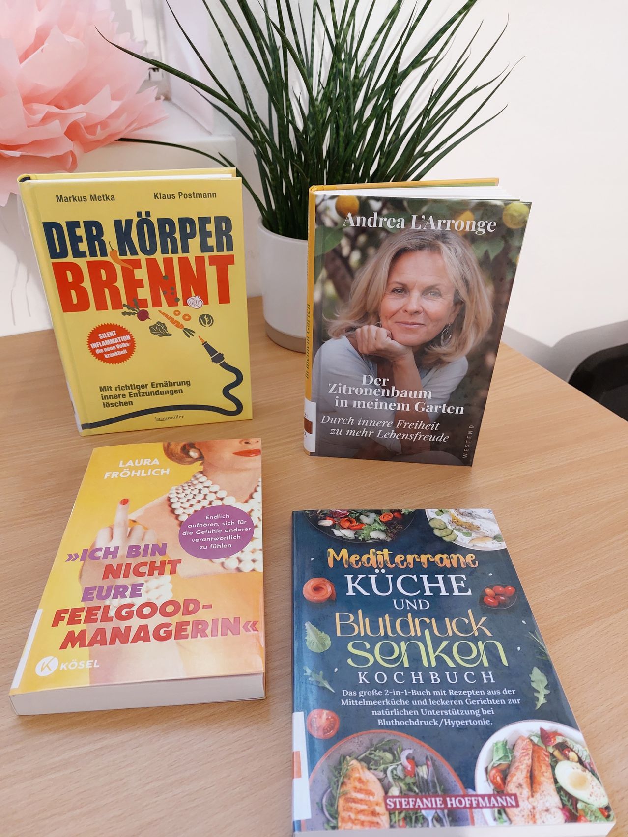Four books on a table: 'Der Körper brennt', 'Der Zitronenbaum in meinem Garten', 'Ich bin nicht eure Feelgood-Managerin', and 'Mediterrane Küche und Blutdruck Senken.'