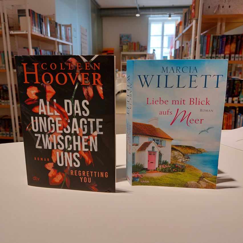 Zwei Romane stehen auf einem Tisch in einer Buchhandlung. Der Buch links ist von Colleen Hoover und heißt 'Alles, was wir nie gesagt haben'. Das Buch rechts ist von Marcia Willett und heißt 'Liebe mit Blick aufs Meer'.