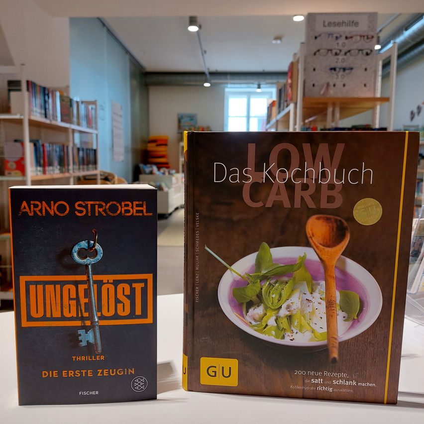 Zwei Bücher stehen auf einem Tisch in einer Bibliothek. Das Buch auf der linken Seite heißt 'Ungelost' von Arno Strobel. Das Buch auf der rechten Seite heißt 'Low Carb' von GU.