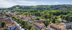 Luftaufnahme einer Stadt mit mehreren Gebäuden, Straßen und üppigen grünen Bäumen unter einem klaren blauen Himmel.