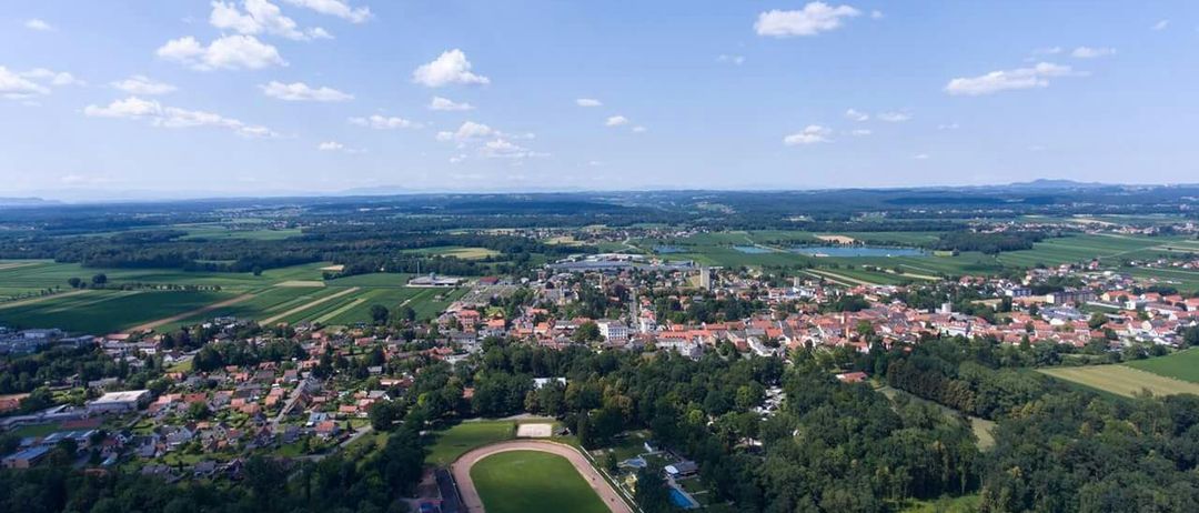 Luftaufnahme einer Stadt mit einem Sportplatz, umgeben von Bäumen unter einem blauen Himmel mit verstreuten Wolken.