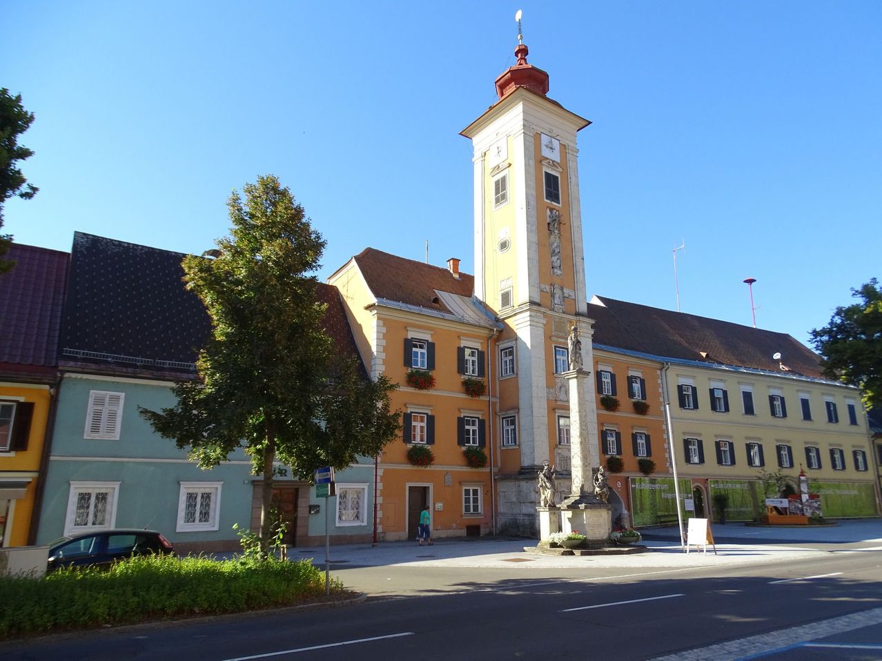Ein hoher Glockenturm steht neben einem Gebäude mit vielen Fenstern und Balkonen. Davor steht eine Statue auf einem Sockel. Eine Person geht auf dem Bürgersteig.