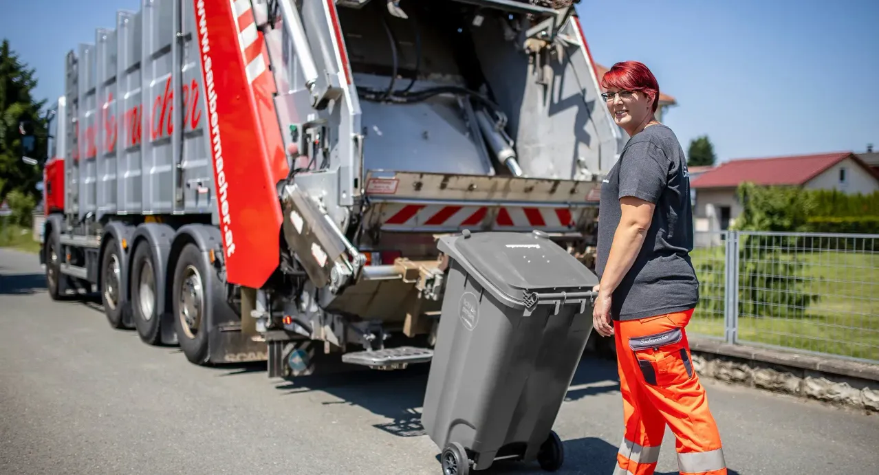 Eine Frau mit roten Haaren, die eine Brille trägt und orange Arbeitskleidung anhat, steht neben einem großen grauen Mülleimer, mit einem roten und weißen Müllwagen im Hintergrund.