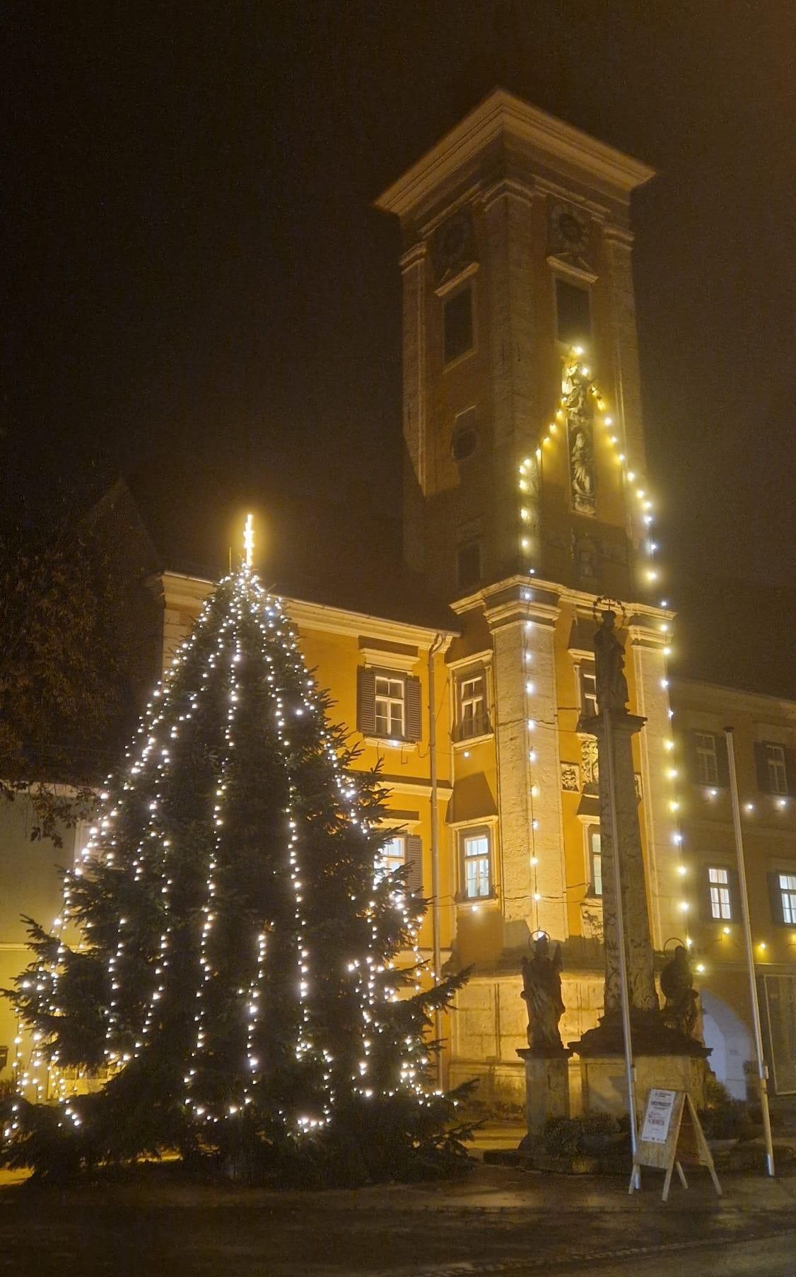 Ein großer beleuchteter Weihnachtsbaum steht vor einem gelben Gebäude mit Statuen, beleuchtet von weißen Lichtern unter einem dunklen Himmel.