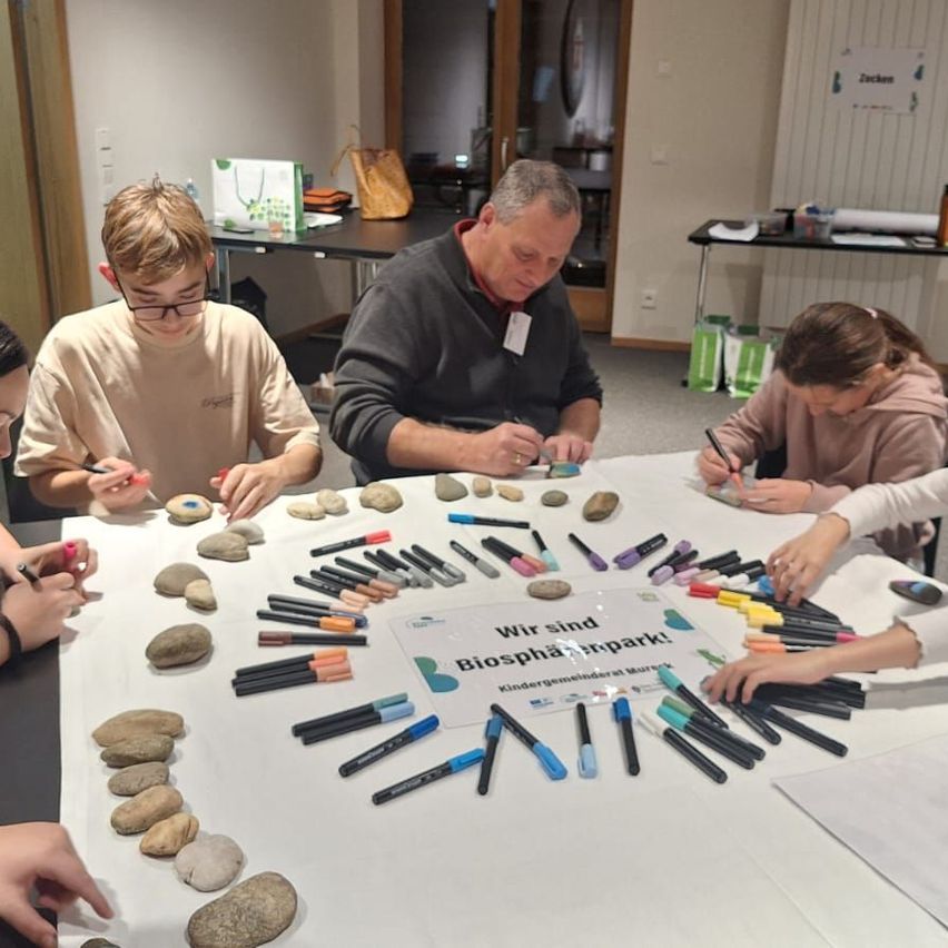 Mehrere Personen arbeiten an einem Tisch mit Steinen und Markern. Eine Person schreibt auf ein Schild, das Wir sind Biosphärenpark! heißt.