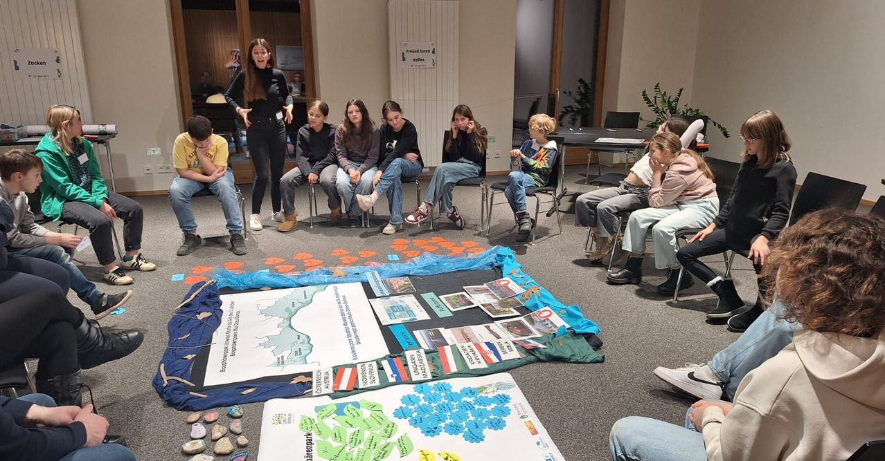 Eine Gruppe von Kindern sitzt um eine große Karte herum, eine erwachsene Frau steht in der Nähe. Papiere und Steine sind auf dem Boden verstreut. Ein Whiteboard mit Text und Bildern steht vor ihnen.
