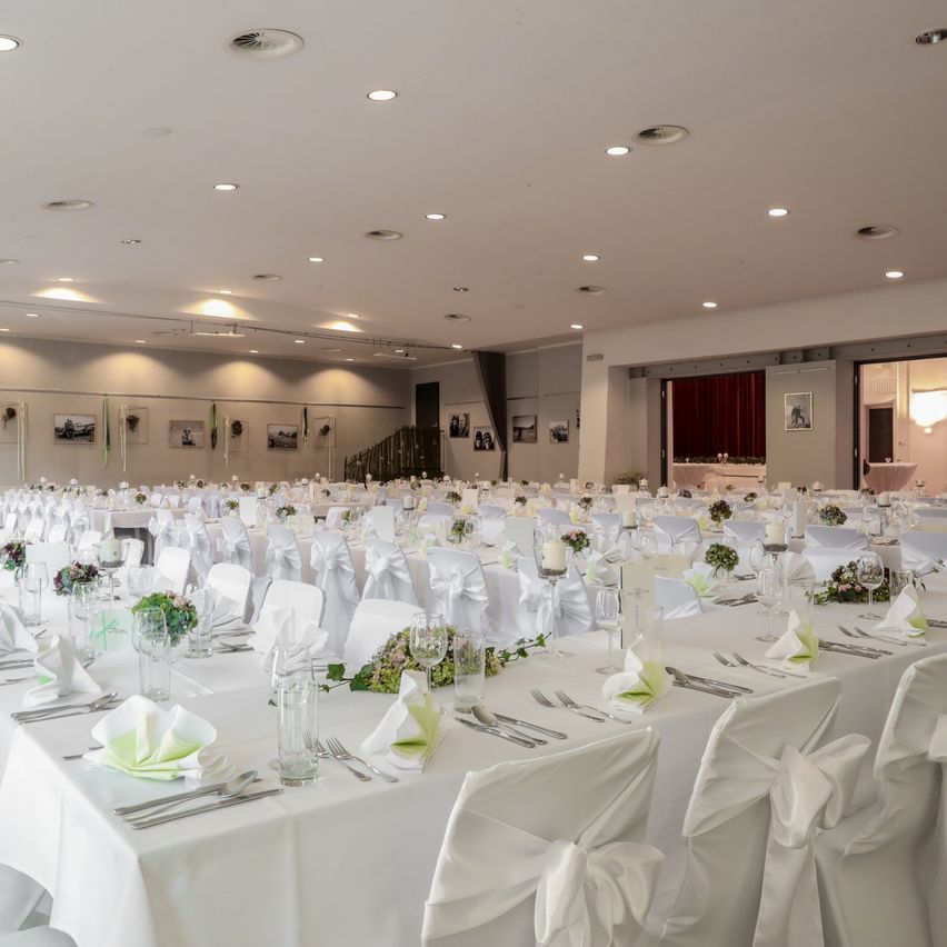 Ein großer Bankettsaal mit mehreren langen Tischen, die für eine Veranstaltung gedeckt sind. Jeder Tisch ist mit weißen Tischdecken bedeckt und die Stühle sind ordentlich aufgestellt. Dekoriert mit grünen Blumenarrangements, weißen Servietten und Besteck.