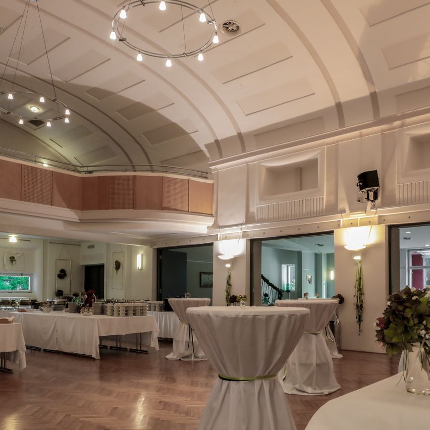 Ein elegant eingerichteter Bankettsaal mit mehreren runden Tischen, die mit Weiß bedeckt sind, einem Holzboden und einer hohen Decke mit dekorativer Beleuchtung.