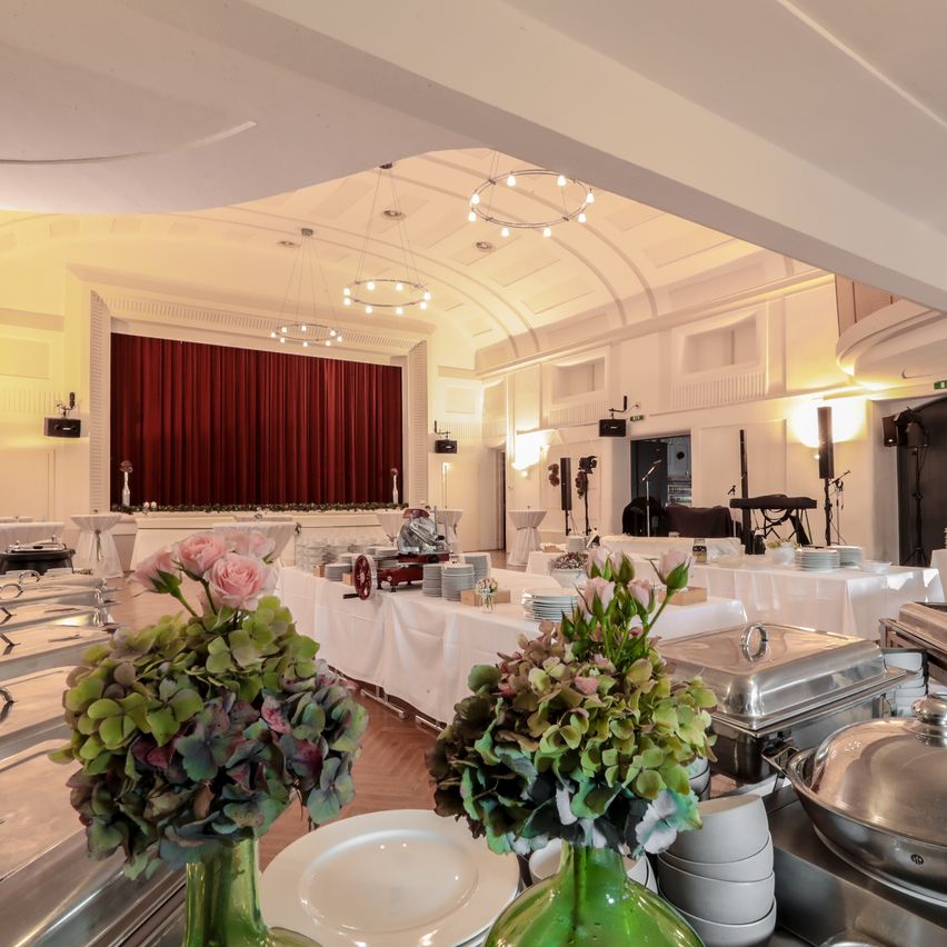 Ein Bankettsaal mit weißen Wänden und einem roten Bühnenvorhang im Hintergrund. Tische sind mit weißen Tischdecken, Blumen, Tellern und Speisen in Chafing-Dishes gedeckt. Eine Bühne ist am Ende des Raumes sichtbar.