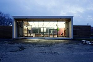 Ein modernes Gebäude mit Glaswänden und -türen, bei Nacht beleuchtet, steht in einem offenen Bereich. Bäume und ein bewölkter Himmel sind im Hintergrund zu sehen.