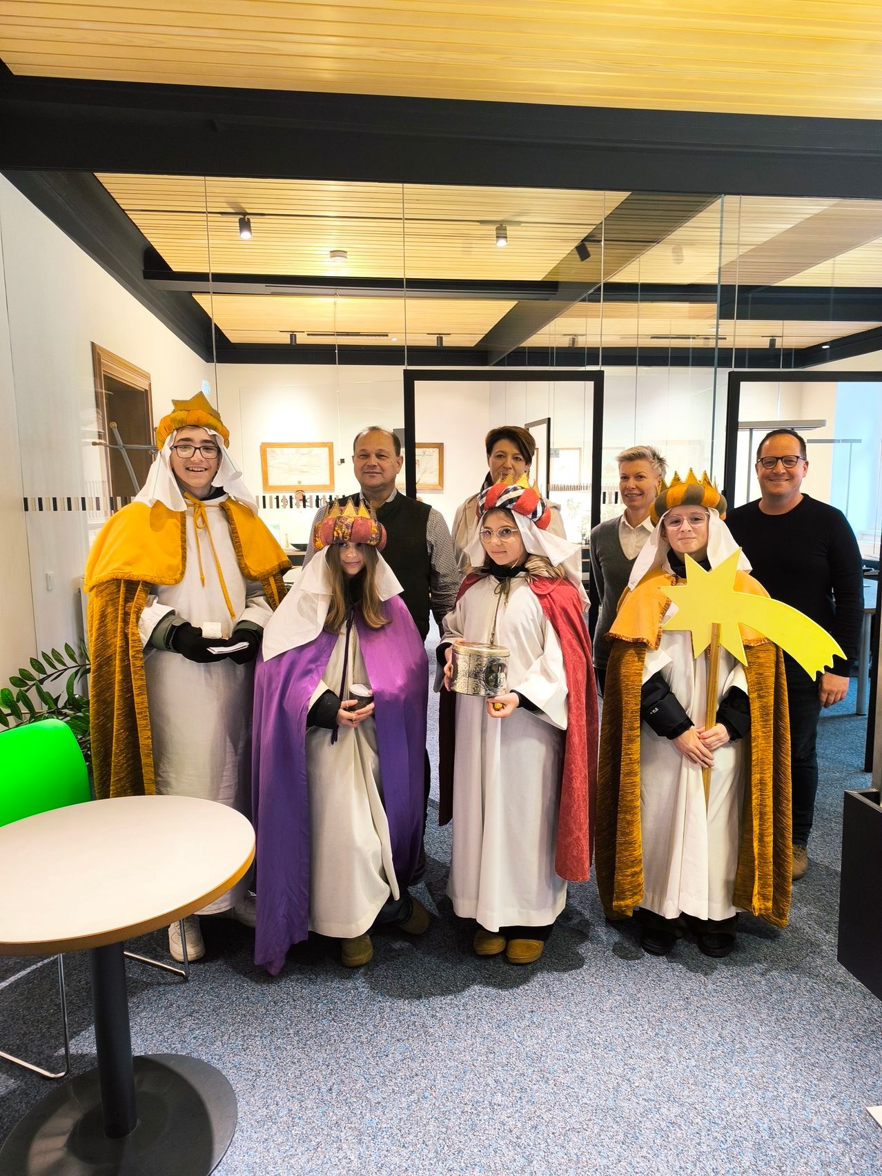 Eine Gruppe von Menschen in Kostümen posiert für ein Foto in einem Raum mit Glaswänden. Es sind zwei Männer und fünf Frauen, eine hält einen Becher und die andere einen Stern.