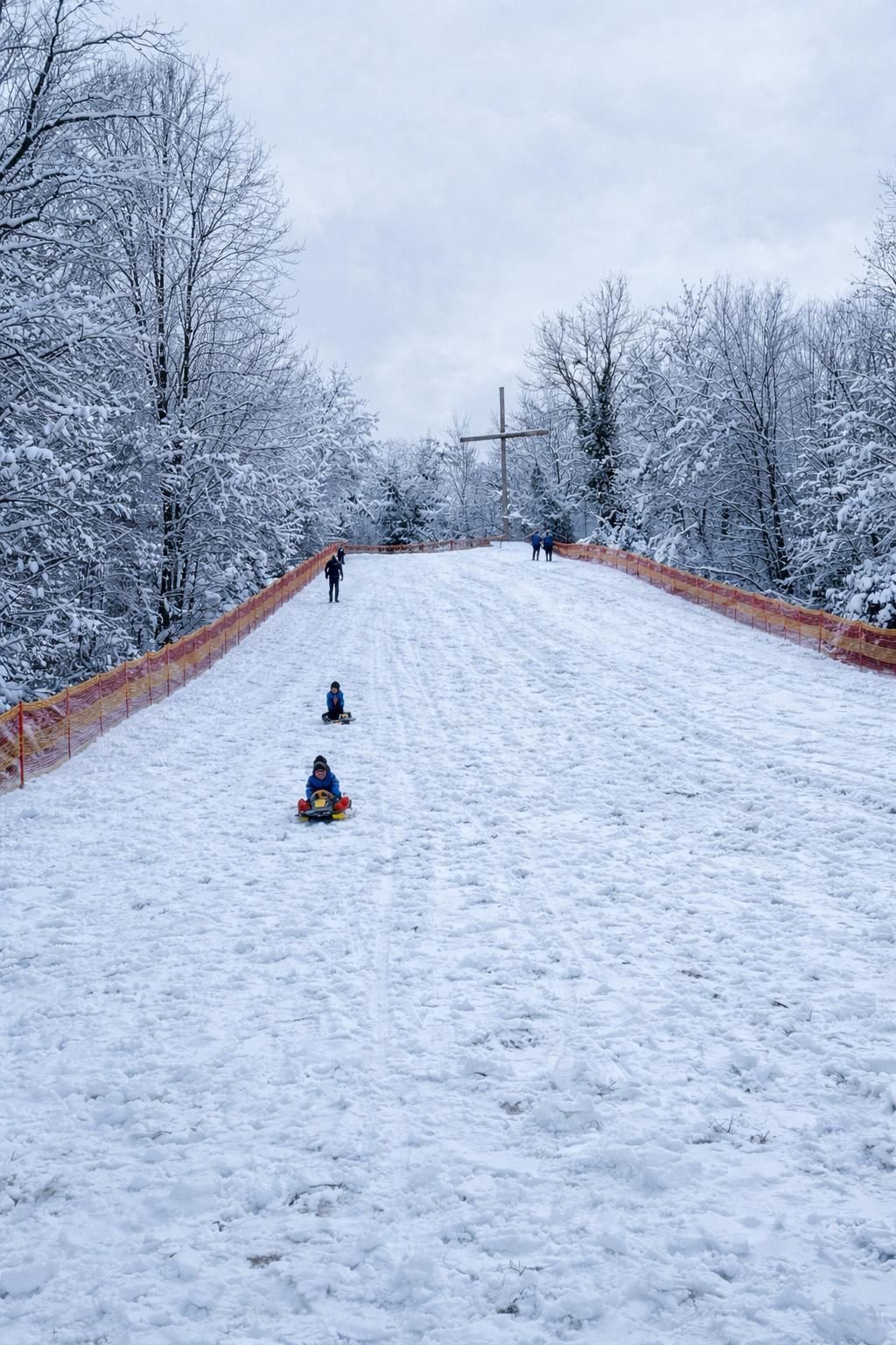 Zwei Kinder rodeln einen schneebedeckten Hügel hinunter, während andere in der Nähe gehen. Im Hintergrund sind schneebedeckte Bäume und ein Kreuz auf einem Hügel zu sehen.