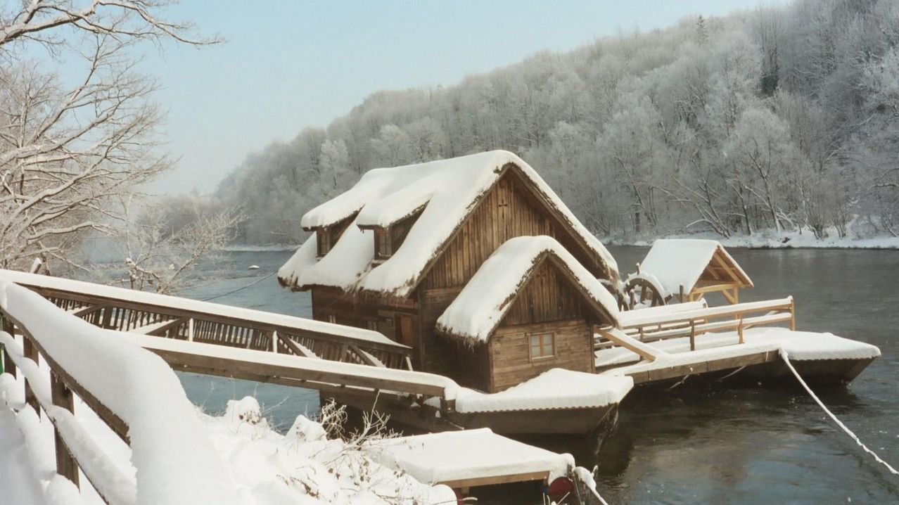 Ein holzverkleidetes Haus auf einem zugefrorenen See, bedeckt mit Schnee, mit einem Steg und einem kleinen Boot in der Nähe.