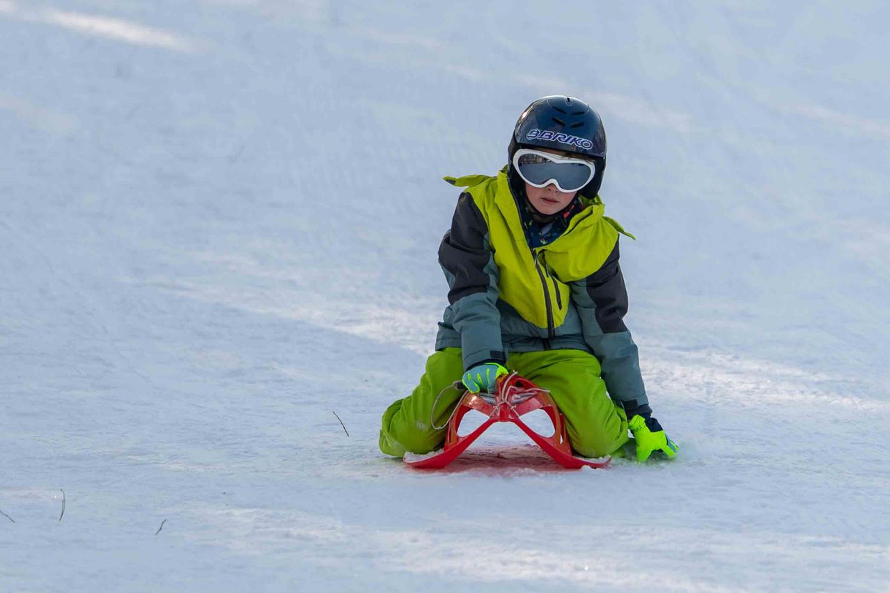 Ein Kind in einer grünen und schwarzen Jacke und einem schwarzen Helm fährt auf einem schneebedeckten Hang mit einem roten Schlitten.