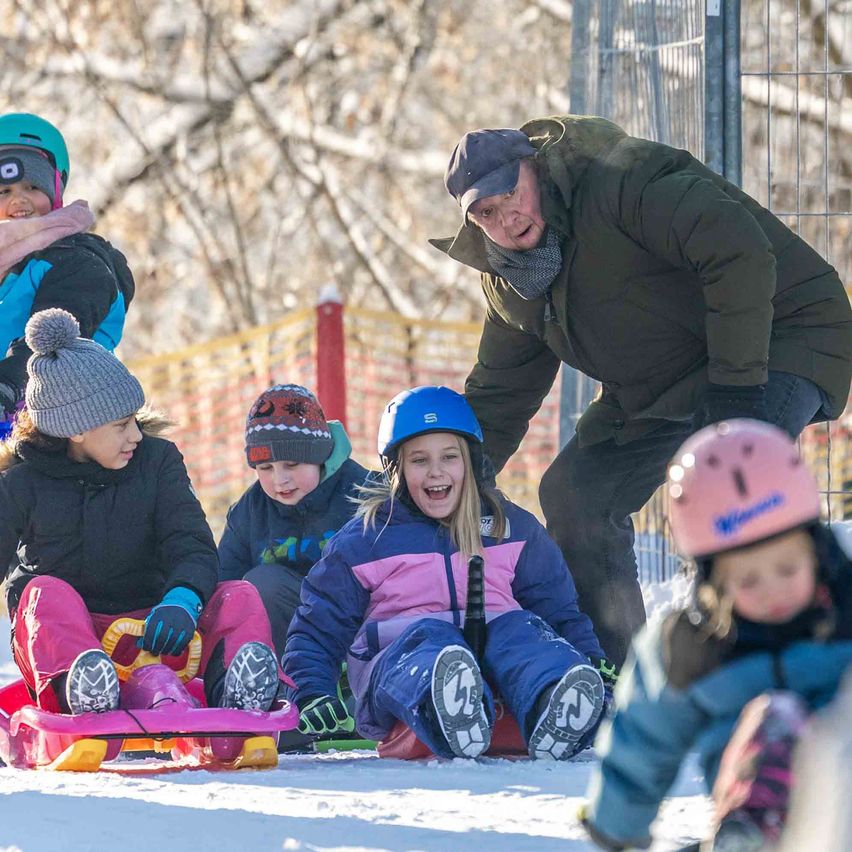 Kinder in Winterkleidung genießen das Schlittenfahren einen schneebedeckten Hügel hinunter, wobei ein Mann ihnen hilft.