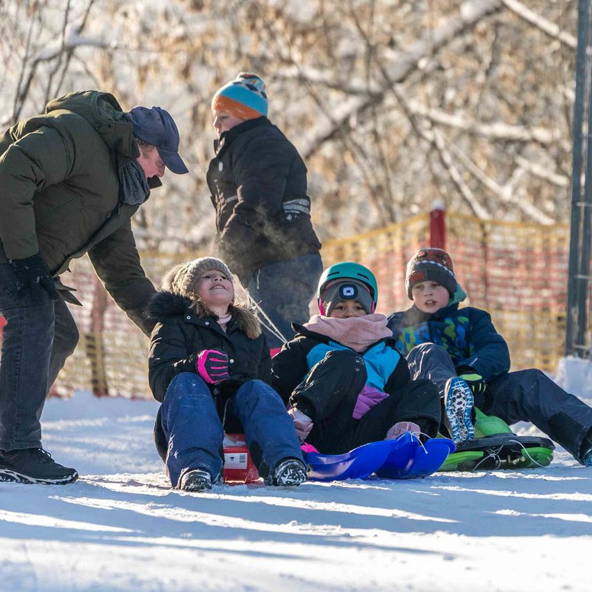 Ein Mann hilft einem Mädchen, eine verschneite Hänge hinunterzurutschen, während andere Kinder in der Nähe warten. Im Hintergrund sind schneebedeckte Bäume zu sehen.