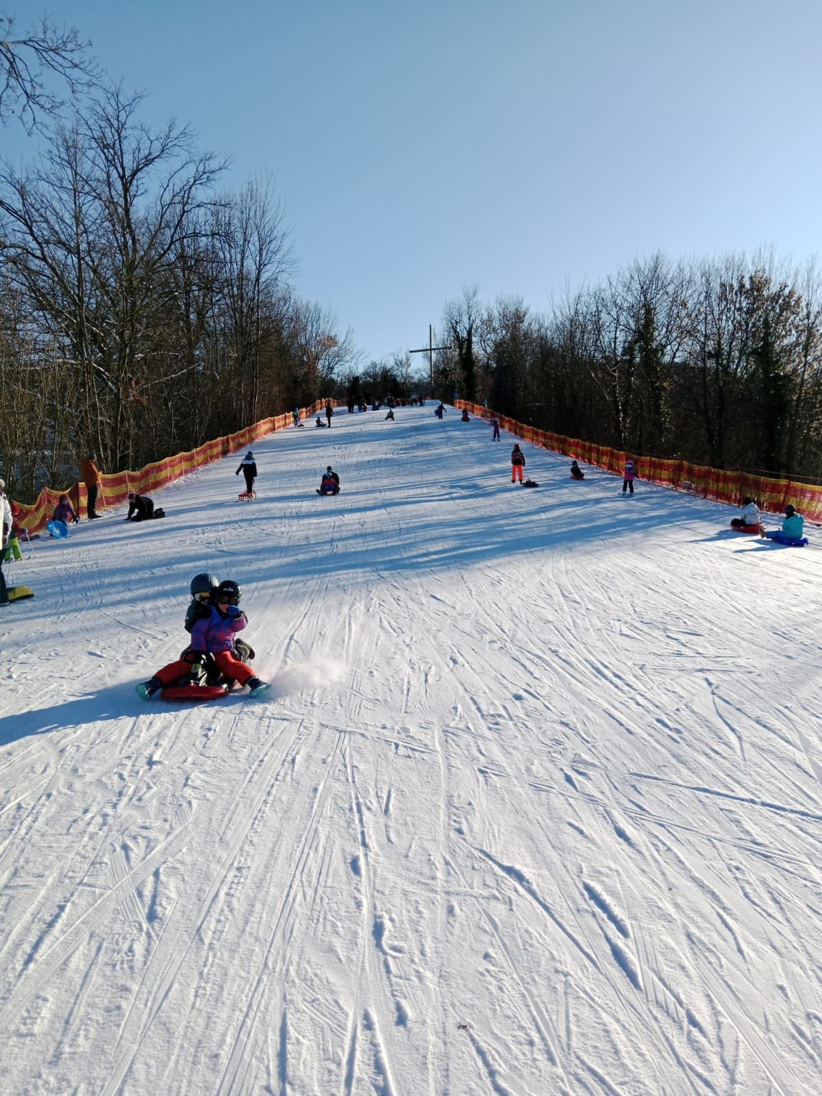 Eine Gruppe von Menschen genießt einen verschneiten Tag auf einer künstlichen Schneerutsche. Einige sitzen, andere stehen. Der Bereich ist mit orange-gelben Barrieren eingezäunt. Bäume umgeben das Gebiet.