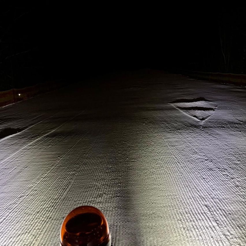 Ein Nachtblick auf eine verschneite Oberfläche, bei der ein rotes Licht am Boden einen kleinen Bereich beleuchtet, Reifenspuren und ein rautenförmiges Loch im Schnee zeigt.