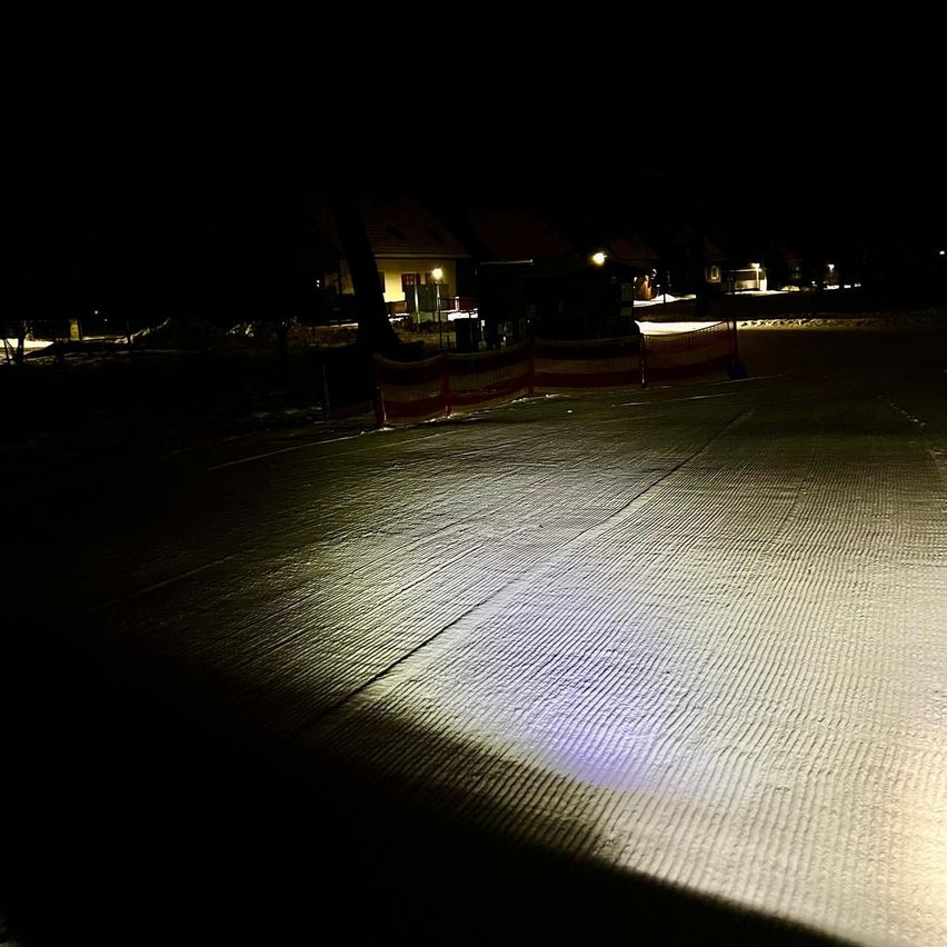 Eine Nachtaufnahme zeigt eine hell beleuchtete Skipiste mit Barrieren und einem Gebäude in der Ferne.