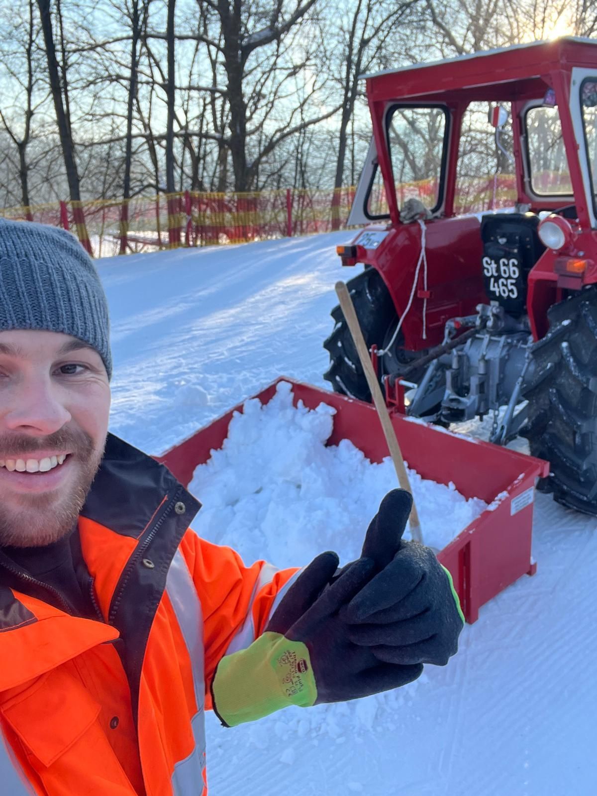 Ein Mann steht lächelnd im Schnee und hält eine Schneeschaufel neben einem roten Traktor mit dem Kennzeichen St 66 465. Bäume und ein Zaun sind im Hintergrund zu sehen.