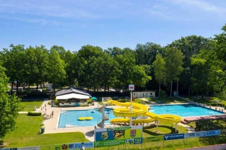 Ein Luftbild eines Wasserparks mit einer großen gelben Wasserrutsche und einem Schwimmbecken, umgeben von üppigem grünem Baumbestand und einem klaren blauen Himmel.
