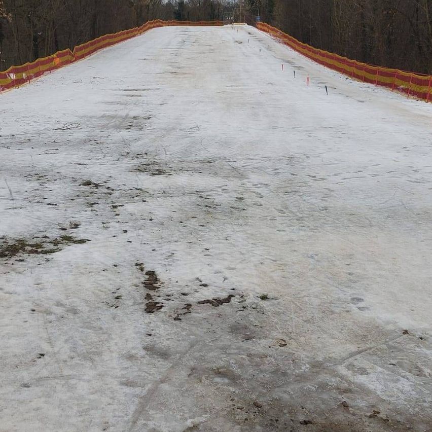 Ein schneebedeckter Hang mit einem roten und gelben Zaun begrenzt ihn. Der Schnee ist uneben mit einigen Fußspuren und Schmutzflecken. Bäume umgeben das Gebiet.