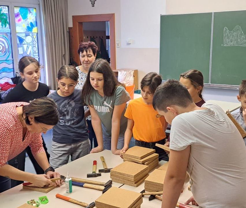 Eine Gruppe von Kindern und Erwachsenen arbeitet in einem Klassenzimmer an einem Projekt, mit Hämmern und Holzplatten. Eine Frau gibt ihnen Anweisungen.
