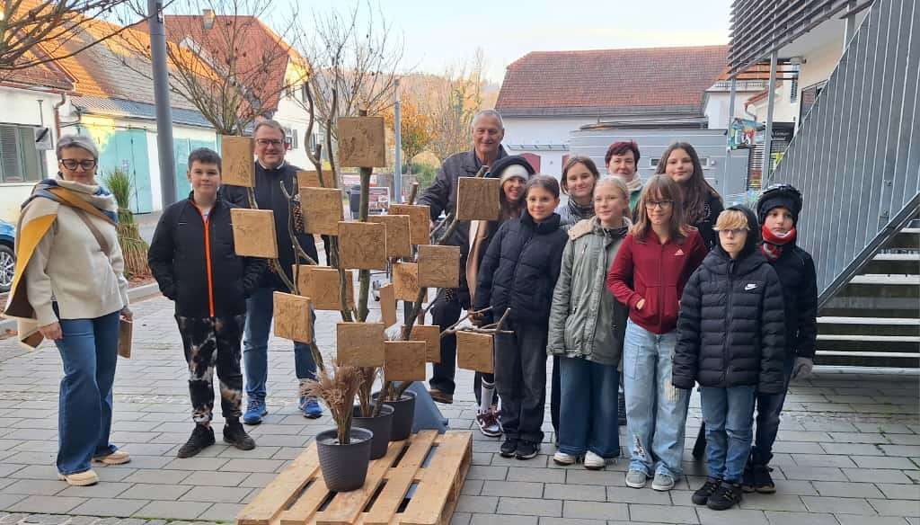 Eine Gruppe von Menschen steht um einen Baum aus Holzblöcken. Sie alle lächeln. Hinter ihnen stehen Häuser und ein Gebäude.