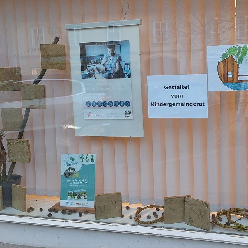 Ein Schaufenster zeigt eine Holzskulptur, ein Bild einer Frau beim Kochen und ein Schild mit der Aufschrift 'Gestaltet vom Kindergemeinderat'. Darunter befindet sich ein Plakat mit 'Biosphärenpark' und anderem Text.