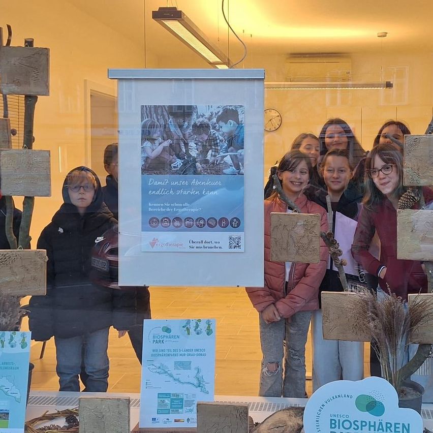 Eine Gruppe junger Mädchen mit lächelnden Gesichtern posiert für ein Foto in einem Raum. Ein Plakat mit Text und QR-Code ist an der Wand befestigt. Einige Holzbretter sind an der Wand platziert. Ein Topf mit Pflanze steht rechts. Eine Person mit Helm steht neben dem Plakat.