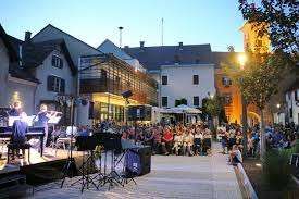 A musical performance is happening in an outdoor area. A few people are sitting on the ground, while others are standing on the sides. A few musical instruments are placed on the stage.