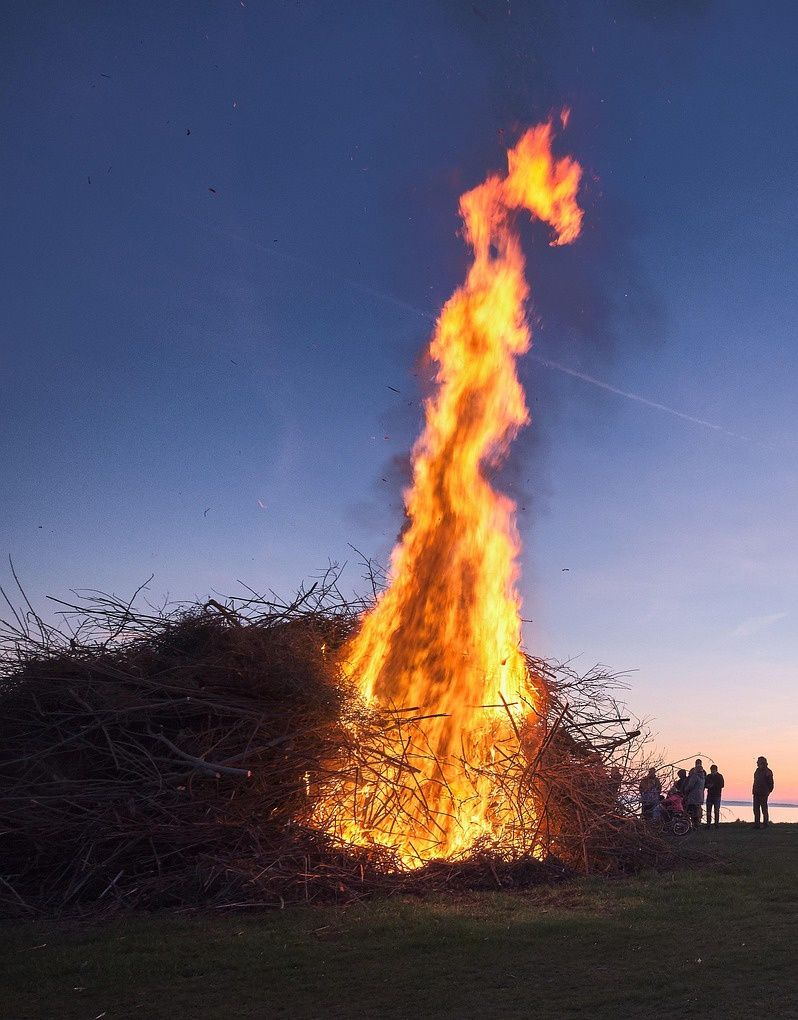 A bonfire burns brightly at dusk, with people gathered around. The fire is surrounded by branches, and the sky is blue with a hint of pink.