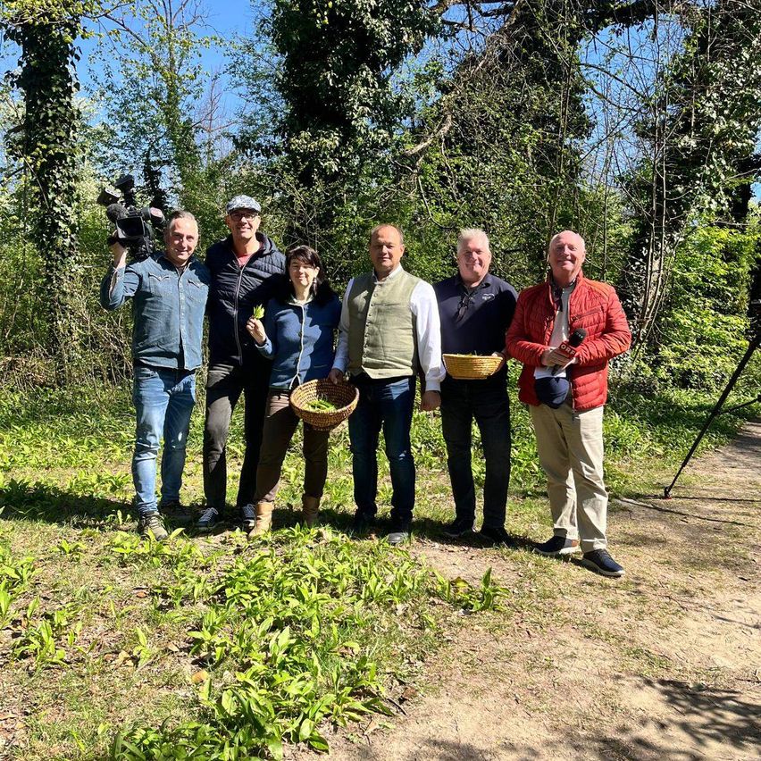 A group of people stands in a grassy area holding baskets. A man on the left holds a camera. They are surrounded by trees and plants.