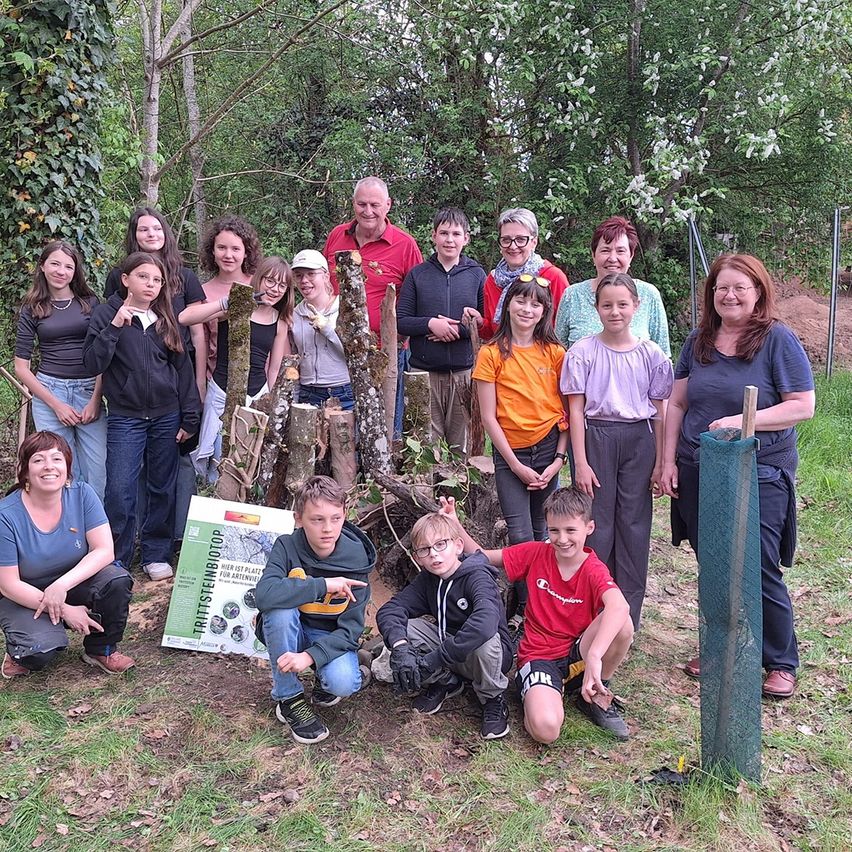 Eine Gruppe von Menschen posiert für ein Foto in einem Wald mit einigen Pflanzen und Bäumen im Hintergrund. Sie alle lächeln und halten einen Holzstab.