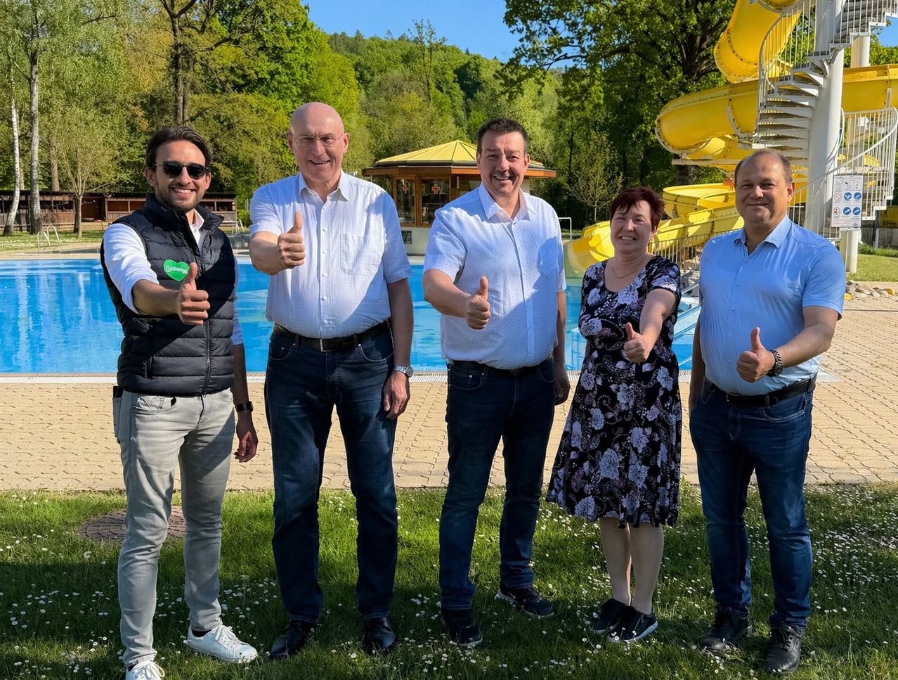 Fünf Personen posieren für ein Foto in einem Park mit einem Pool und Wasserrutschen im Hintergrund. Sie alle haben Lächeln und Daumen hoch.