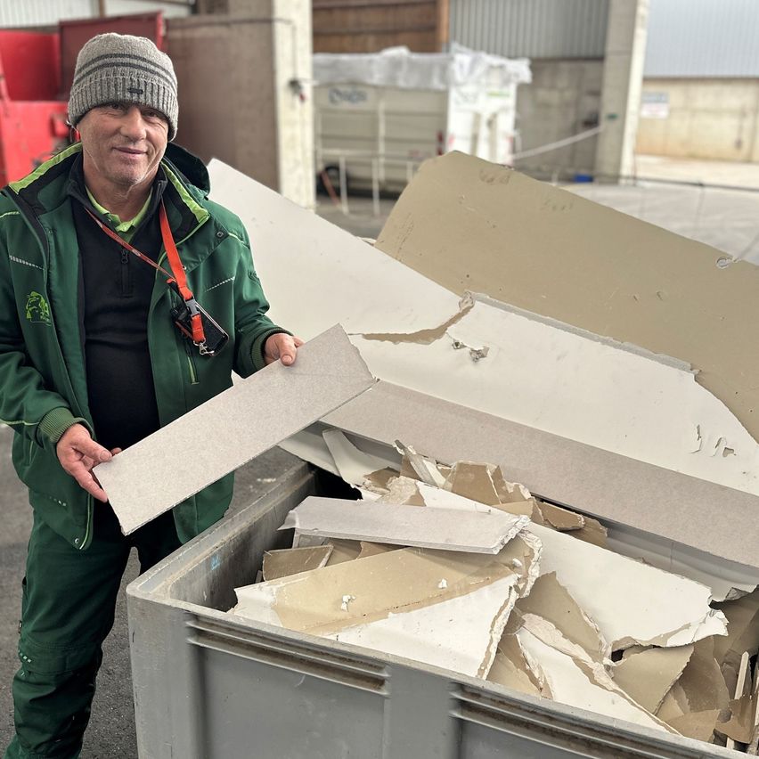 Ein Mann in einer grünen Jacke hält ein Stück Pappe inmitten eines Haufens weggeworfener Materialien im Freien.