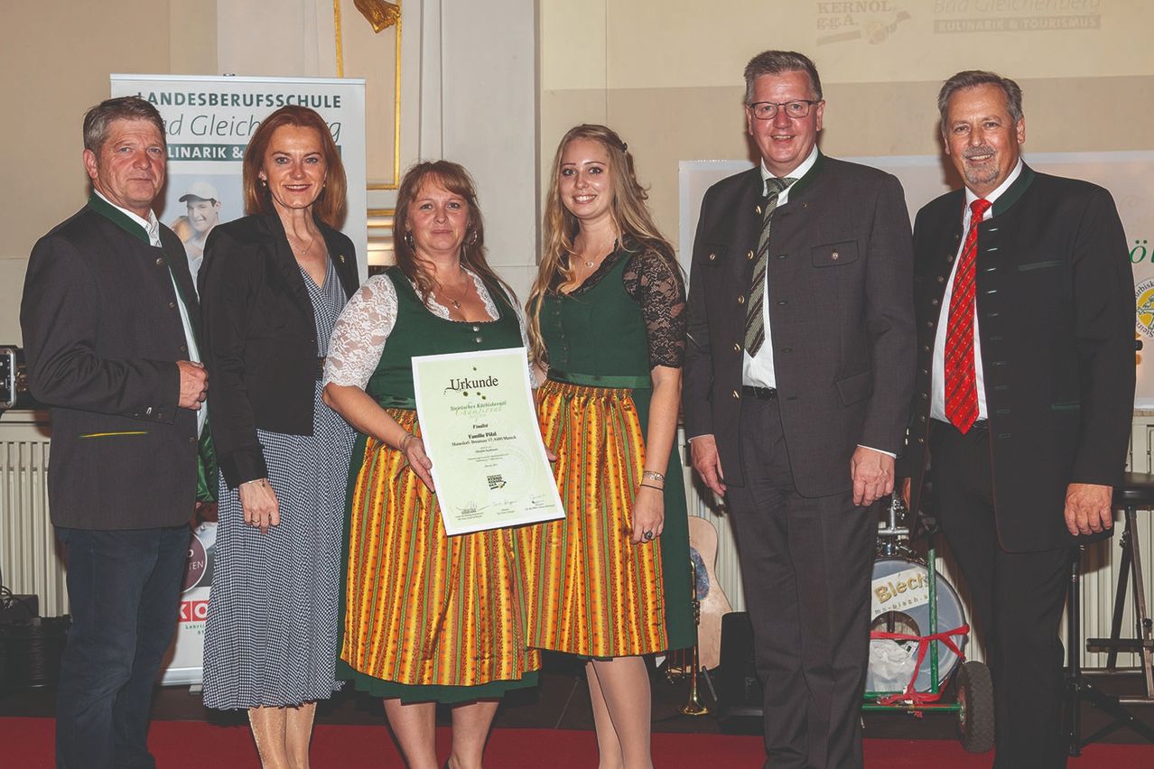 Zwei Frauen in traditioneller deutscher Kleidung stehen mit einem Zertifikat und zwei Männern, einer mit Brille und Krawatte. Dahinter ein Hintergrund mit dem Text 'Gleich' und ein Schild. Eine Trommel und eine Trompete liegen auf dem Boden.